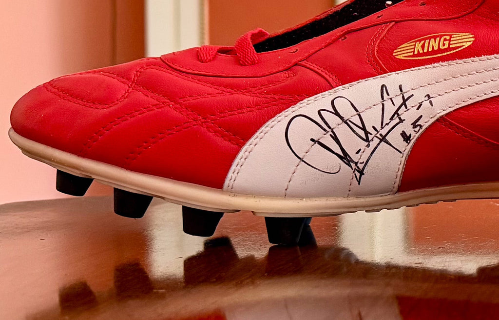 A pair of red Puma soccer cleats with white stripes and autographed signatures, displayed on a reflective wooden table in an indoor setting.