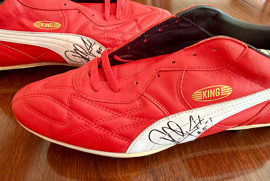 A pair of red leather Puma King soccer cleats with white side stripes and black autographs, resting on a wooden floor.