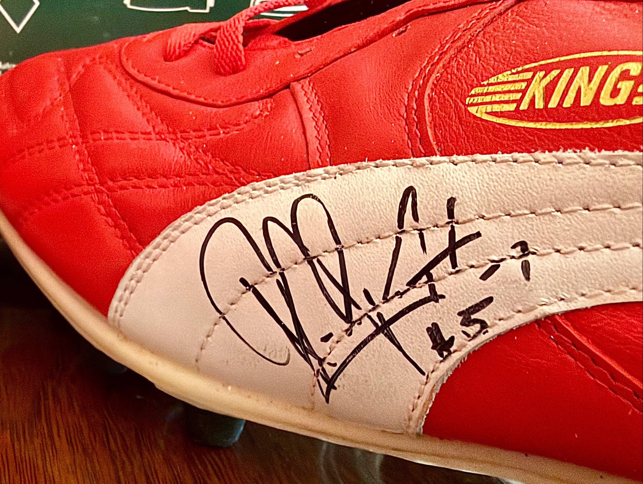 Two red and white soccer cleats signed in black marker, displayed side-by-side on a wooden surface with a green shoe sizing chart in the background.